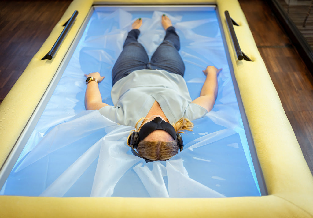 Person lying fully clothed on a blue-lit Driifloat membrane, wearing headphones and an eye mask.