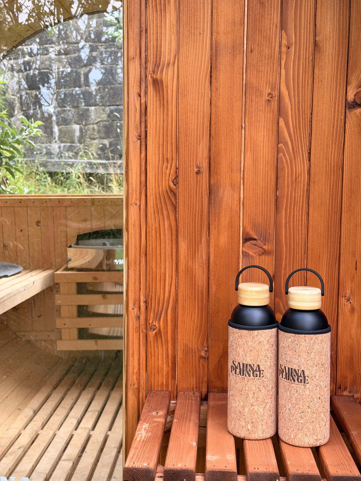 Two cork-covered SaunaPlunge bottles beside a barrel sauna doorway